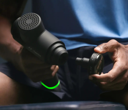 A person holding a Hyperice Hypervolt 2 percussion massage gun, attaching a massage head attachment. The device has a sleek black design with ventilation holes and a glowing green ring near the handle, indicating power status. The user is seated, wearing a blue shirt and dark shorts, preparing the massage gun for use.