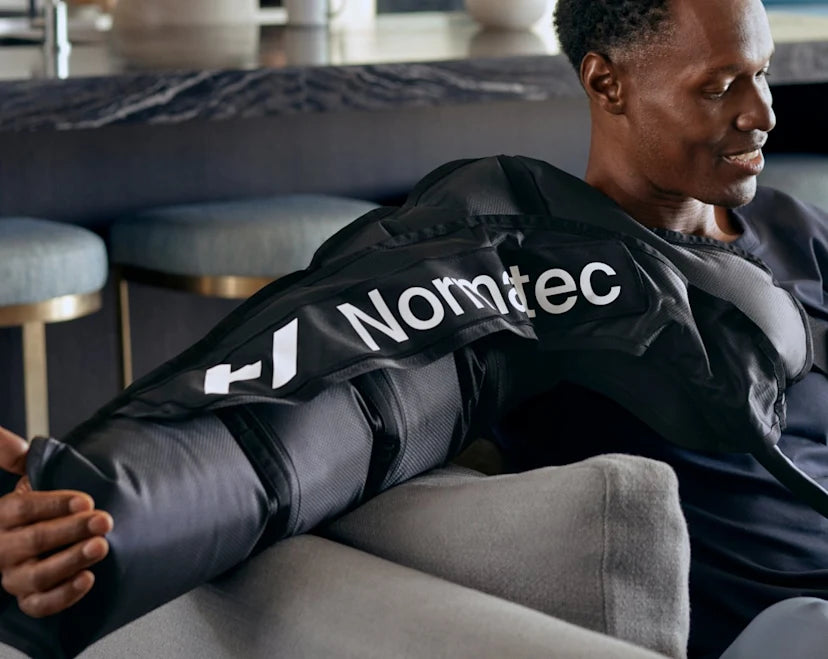 A man sitting comfortably on a couch using the Normatec 3 arm attachment for compression therapy. The black, high-tech sleeve with 'Normatec' branding covers his entire arm, secured with adjustable straps. He appears relaxed, enjoying the benefits of dynamic air compression for muscle recovery. The background features a stylish modern interior with bar stools and a marble countertop.