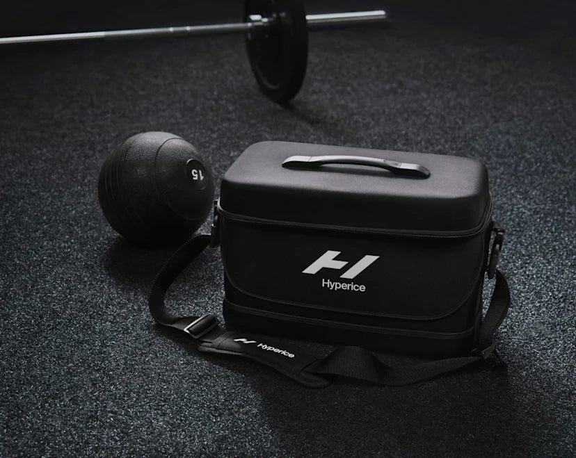 A black Hyperice Normatec carrying case with a sleek, compact design sits on a dark gym floor. The case features a sturdy handle on top, a zippered closure, and an adjustable shoulder strap with the Hyperice logo. Positioned nearby are a black 15-pound medicine ball and a barbell, emphasizing the case’s use in fitness and recovery settings. The lighting is moody, creating a professional and high-performance aesthetic.